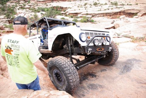 1995 Ford Bronco - A heavily modified white Ford Bronco is parked on a rocky, desert terrain. A man wearing a light green t-shirt with text and a black baseba…