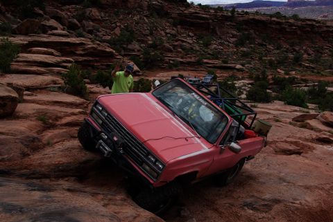 1995 Ford Bronco - A pink off-road modified pickup truck navigates a rocky, uneven terrain. A person in a lime green shirt stands on a rock formation in the b…