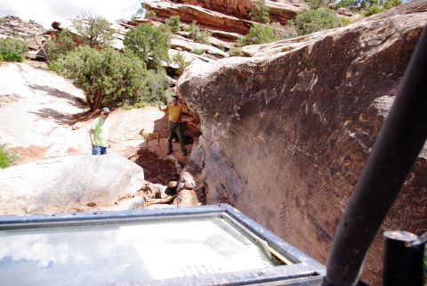 1995 Ford Bronco - Two men and a dog stand near a large rock formation in a desert landscape. The foreground shows part of a vehicle's interior, including a w…