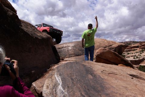 1995 Ford Bronco - A red off-road vehicle is navigating a rocky incline with a person in a green shirt waving in the foreground. Another person is taking a ph…