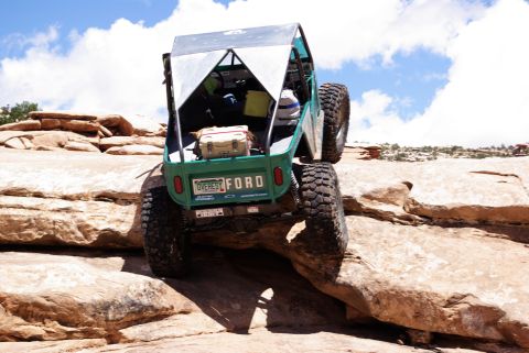 1995 Ford Bronco - The rear view of a green custom off-road vehicle with large tires is navigating a rocky, desert terrain under a blue sky with white clouds.…