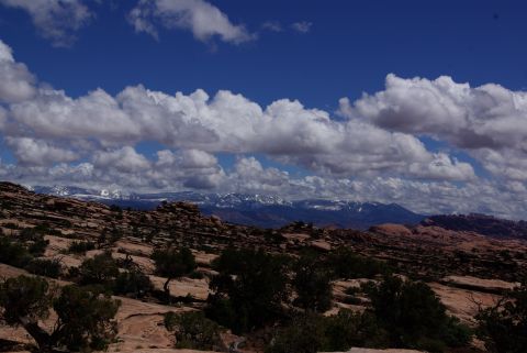 1995 Ford Bronco - A wide landscape shot shows a vast expanse of desert terrain under a bright blue sky filled with dramatic, puffy white clouds. In the dista…