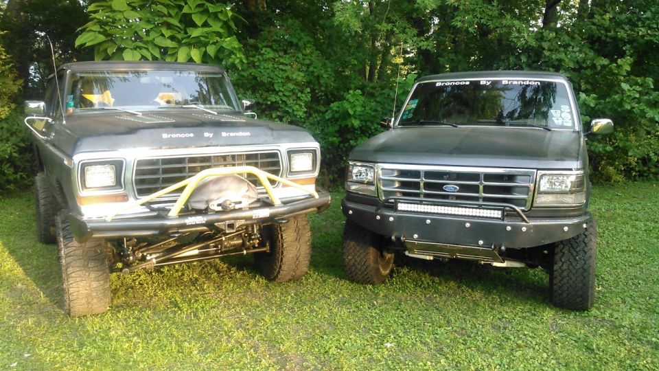 Two Ford Broncos parked on grass, one older with yellow brush guard, one newer with light bar.
