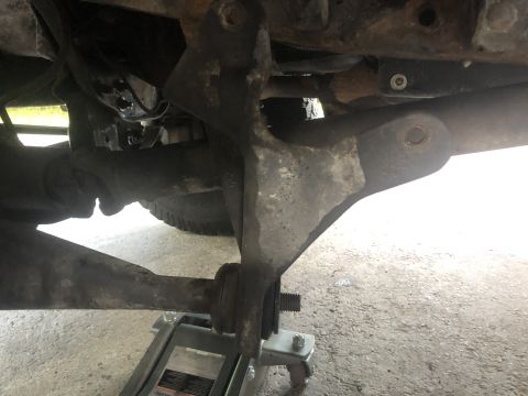 1995 Ford Bronco - The underside of a vehicle shows a suspension component, likely a control arm, supported by a hydraulic jack. The part is heavily weathered…