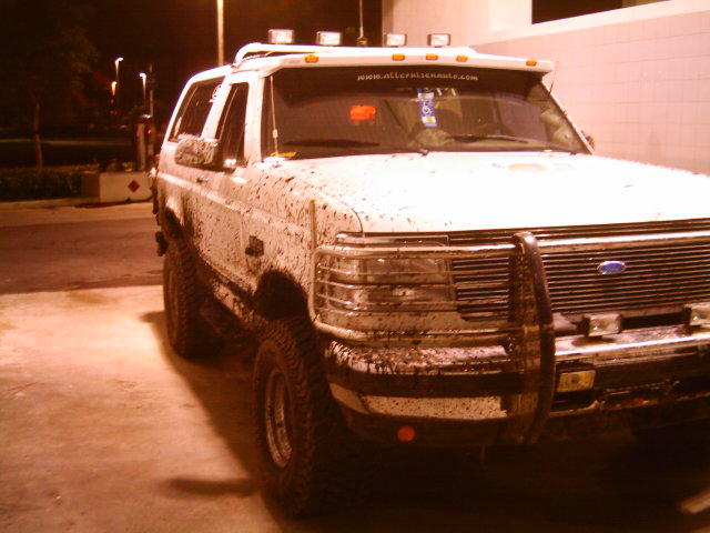 Mud-covered white 1990s Ford Bronco with roof lights and brush guard at night.