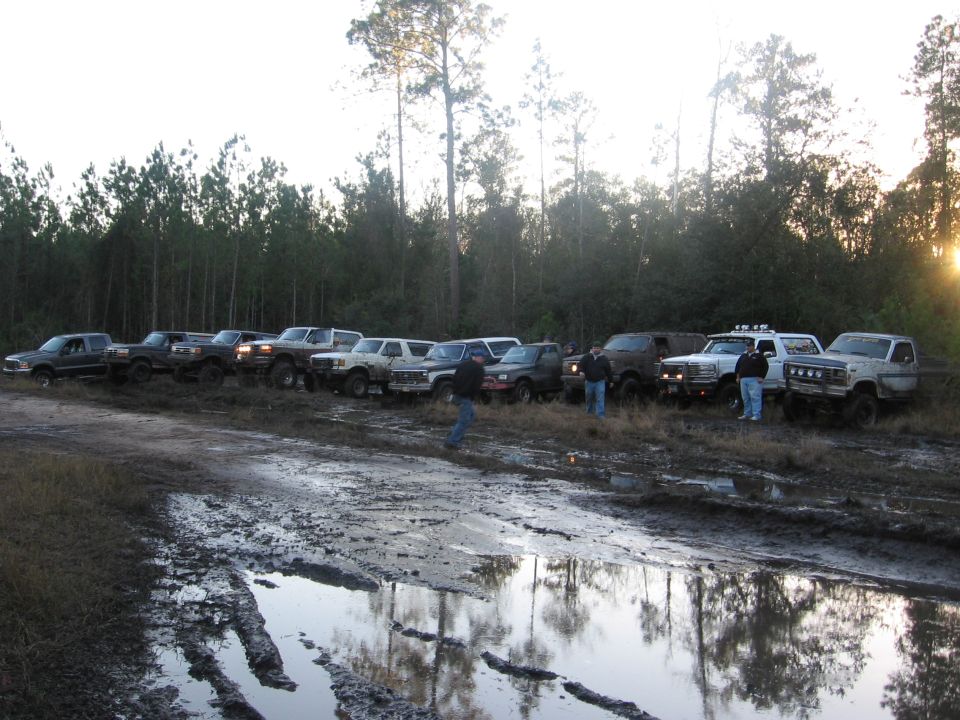 Several pickup trucks and SUVs parked in a muddy area with a forest in the background.