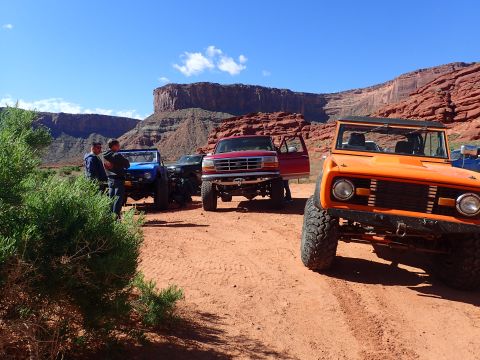 1995 Ford Bronco - A group of off-road vehicles are parked on a dirt road in a desert landscape with red rock cliffs in the background. The vehicle in the for…