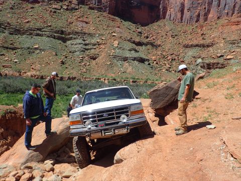 1995 Ford Bronco - A white Ford Bronco is navigating a rocky, off-road trail with several people standing around it. The vehicle is tilted at an angle as it c…