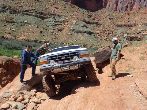 1995 Ford Bronco - A white Ford Bronco is navigating a rocky, off-road trail with three people assisting. The vehicle is angled steeply, with its front tires…
