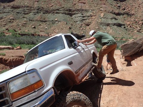 1995 Ford Bronco - A white Ford Bronco is stuck on a rocky, off-road trail with a person pushing on the driver's side door. The vehicle is tilted at an angle…
