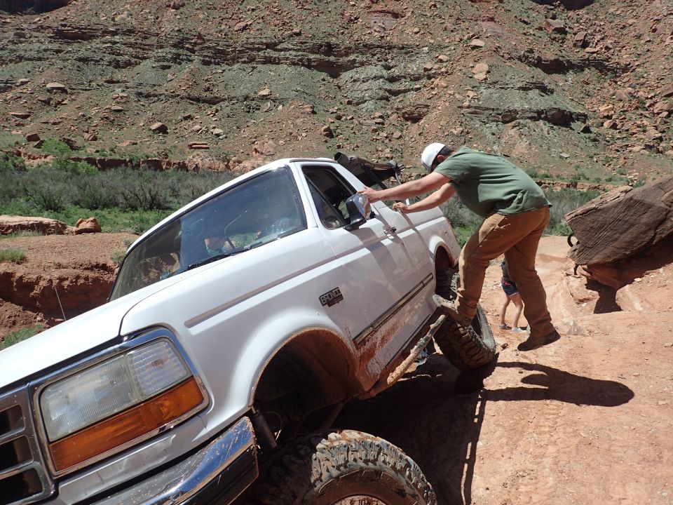 White Ford Bronco stuck on a rocky trail, tilted with front tire off the ground, person pushing door.
