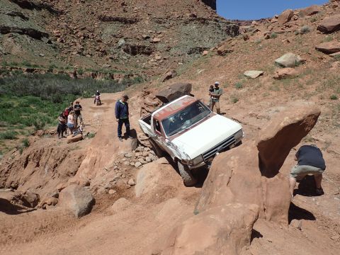 1995 Ford Bronco - A white and red Ford pickup truck is navigating a rocky, off-road trail with several people observing. The truck is tilted at a steep angle…