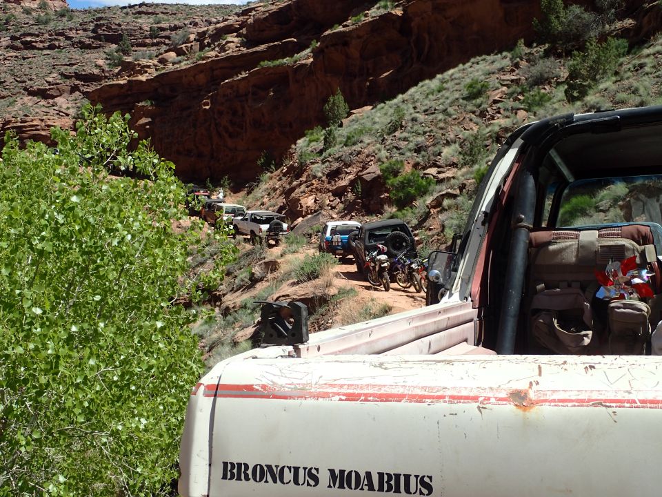Off-road convoy on a dusty canyon trail, foreground shows rear of white pickup truck with text.
