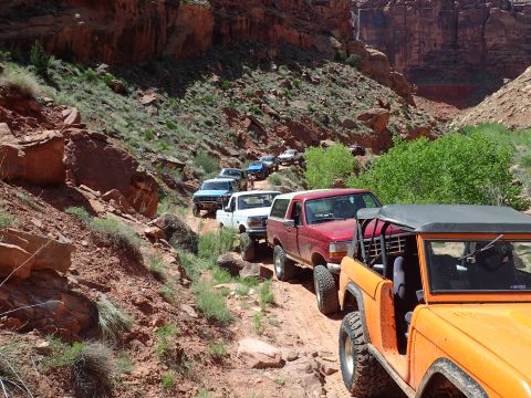 1995 Ford Bronco - A line of off-road vehicles, including a red Ford Bronco and an orange Jeep CJ, navigate a rocky, dirt trail through a canyon with steep re…