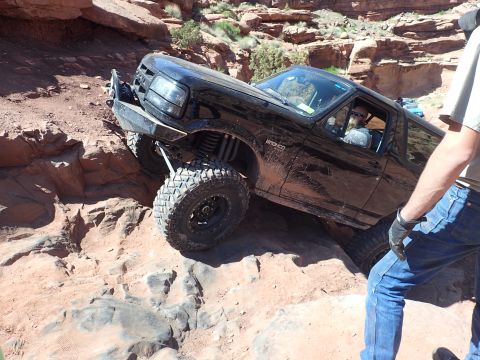 1995 Ford Bronco - A black Ford Bronco is navigating a steep, rocky off-road trail. The vehicle is tilted at a sharp angle as its tires climb over large bould…