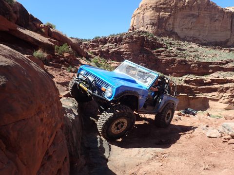 1995 Ford Bronco - A bright blue Ford Bronco is navigating a steep, rocky off-road trail with large boulders and red rock formations surrounding it. The vehic…