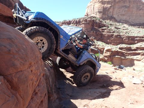 1995 Ford Bronco - A blue Ford Bronco is navigating a steep, rocky incline with its driver behind the wheel. The vehicle is angled precariously, with its fron…