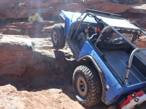 1995 Ford Bronco - A blue off-road vehicle navigates a rocky, dusty trail with a driver at the wheel. The vehicle is angled, showing its large tires and modif…