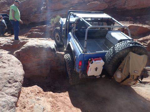 1995 Ford Bronco - A blue off-road vehicle with a roll cage is navigating a rocky, desert terrain. The vehicle has a spare tire mounted on the back, along wit…