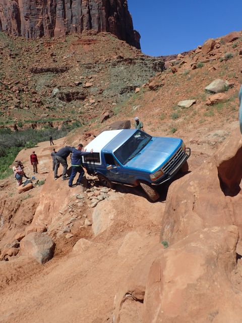 1995 Ford Bronco - A blue Ford Bronco with a white camper shell is navigating a steep, rocky, off-road trail. Several people are gathered around the vehicle,…
