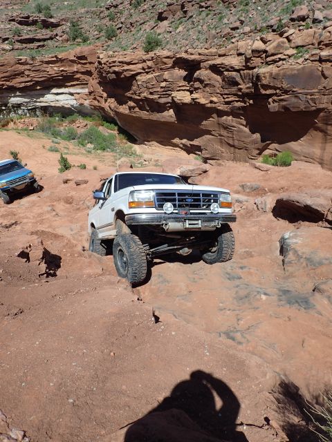 1995 Ford Bronco - A white Ford Bronco is navigating a rocky, off-road trail with steep red rock formations in the background. Another blue vehicle is visible…