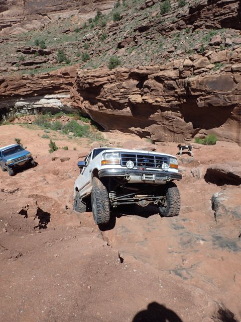 1995 Ford Bronco - A white Ford Bronco is navigating a rocky, off-road trail with large tires and lifted suspension. Another blue SUV is visible further up th…