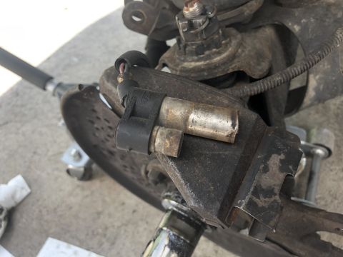 1995 Ford Bronco - Close-up of an automotive cast bracket with a cylindrical solenoid/actuator and connector mounted on a worn metal assembly.