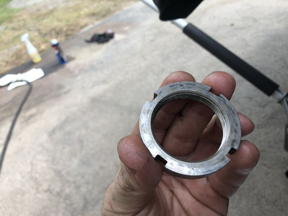 Close-up of a metal locknut with notches, held in a hand. Background shows a pole and cleaning supplies.