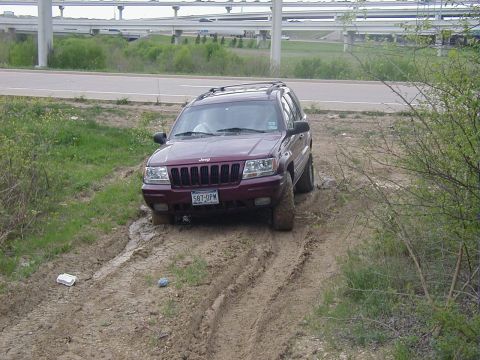 1991 Ford Bronco - Three-quarter front view of a maroon Jeep Grand Cherokee sitting in deep rutted mud on a lot beside a roadway, mud caked on the tires and l…