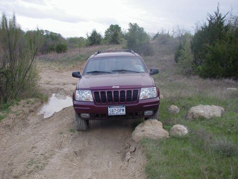 1991 Ford Bronco - Head-on view of a maroon Jeep Grand Cherokee (WJ-generation) sitting on a rutted dirt two-track with one front wheel up on rocks, a muddy p…