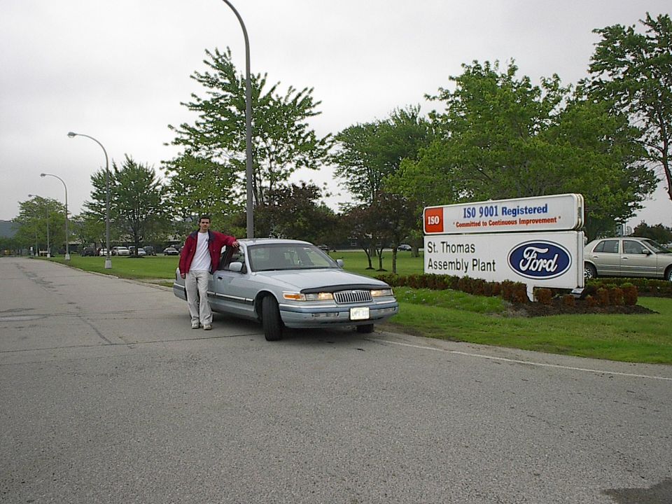 Man leaning on a 1985 Mercury Grand Marquis sedan in front of a Ford Assembly Plant sign.