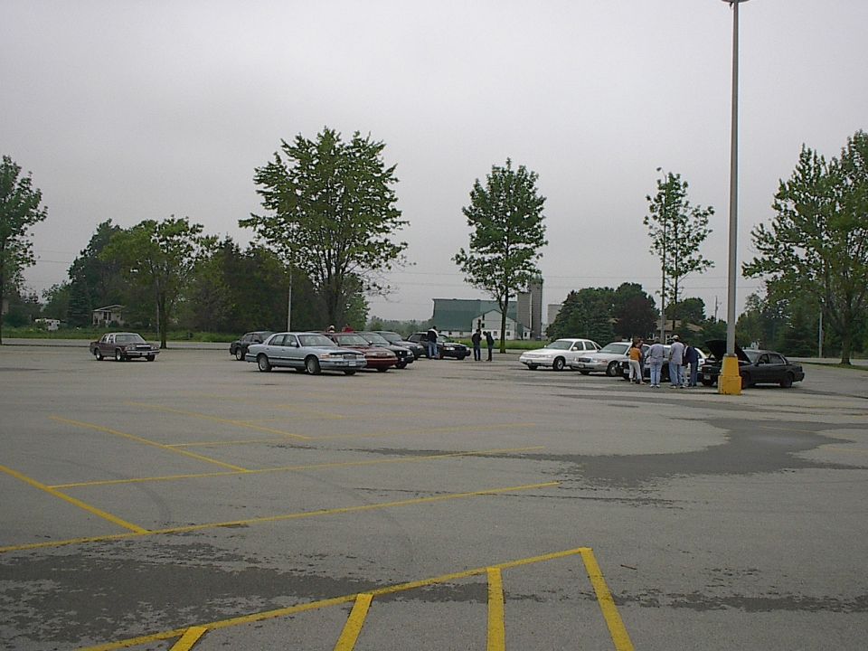 Group of people gathered around cars in a parking lot, one car hood open.