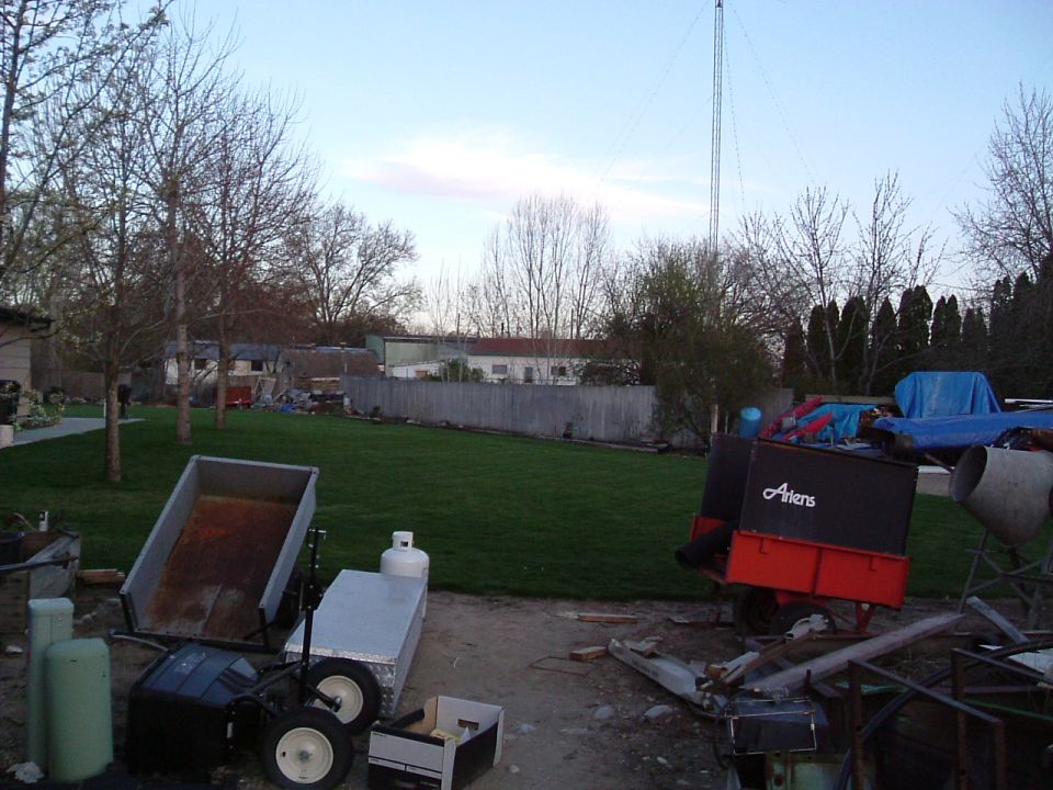 Yard with a dump trailer, propane tank, and a red and black trailer with 'Ariens' logo. Tall tower in background