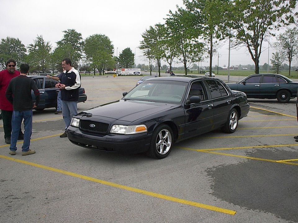 Black Ford Crown Victoria sedan parked in a lot with people around.