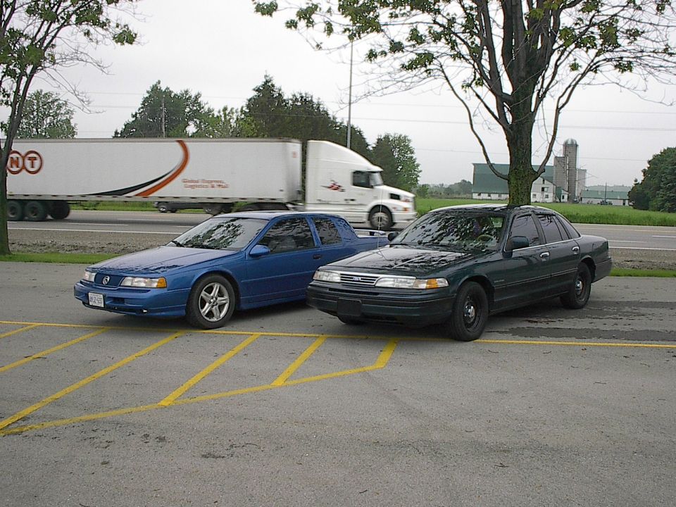 Blue sedan parked next to a dark green sedan in a parking lot with a semi-truck in the background.