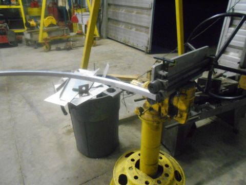 1992 Ford Bronco - A metal bending machine is set up in a workshop with various metal scraps and a large trash bin nearby. A long piece of metal is being fed…