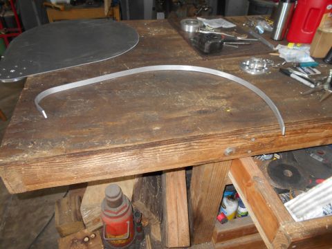 1992 Ford Bronco - A curved piece of metal is laid across a wooden workbench. A large, flat, teardrop-shaped piece of metal with several holes drilled in it i…