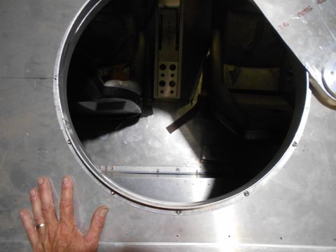1992 Ford Bronco - A hand with a ring is placed on the edge of a large circular opening in a metallic surface, revealing a dark interior with two seats and so…