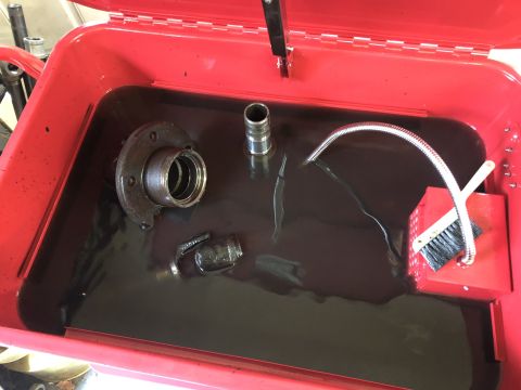 1995 Ford Bronco - A red parts washer tub is filled with dark fluid, containing various metal automotive parts and a brush. The lid of the tub is open, reveal…