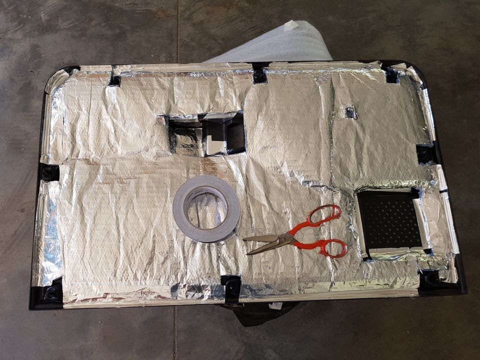 Car door panel covered in silver sound deadening material, with a roll of tape and scissors on top