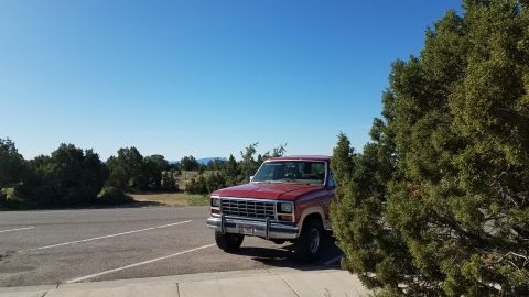 1985 Ford Bronco - A red Ford Bronco is parked on the side of the road with trees and a dry landscape in the background.