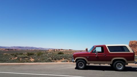 1985 Ford Bronco - A two-tone Ford Bronco sits parked on the side of a road with a vast desert landscape stretching out behind it under a clear blue sky.