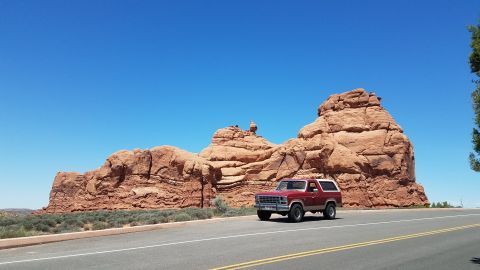 1985 Ford Bronco - A red Ford Bronco drives past large red rock formations under a clear blue sky.