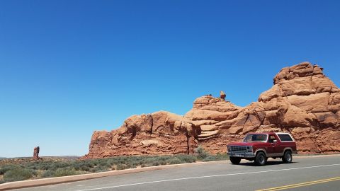 1985 Ford Bronco - A red 1985 Ford Bronco travels along a road with the iconic red rock formations of Arches National Park in the background.