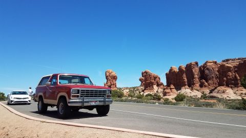 1985 Ford Bronco - A red and tan Ford Bronco drives down a highway with distinctive red rock formations under a clear blue sky.