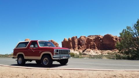 1985 Ford Bronco - A red and tan 1985 Ford Bronco is parked on the side of a road with a scenic desert landscape and rock formations.