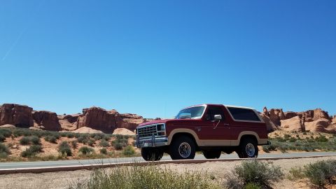 1985 Ford Bronco - Red and tan 1985 Ford Bronco travels through a desert landscape under a clear blue sky.