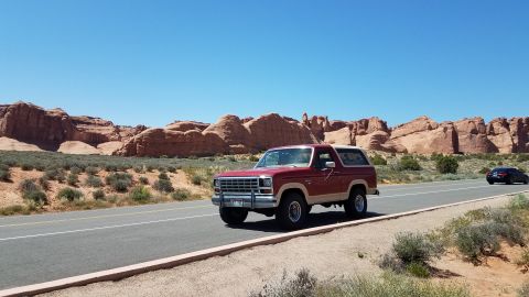 1985 Ford Bronco - A classic Ford Bronco cruises down a desert highway under a clear blue sky.