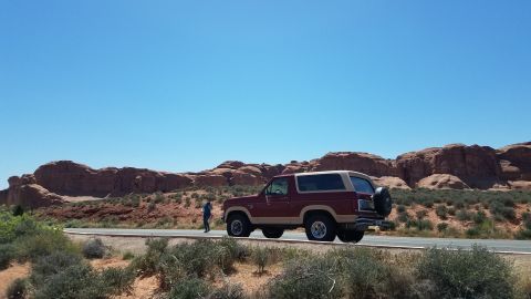 1985 Ford Bronco - A maroon and tan Ford Bronco is parked on the side of a road in a desert landscape with red rock formations.