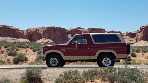 1985 Ford Bronco - A classic Ford Bronco is seen driving through a desert landscape with distinctive rock formations under a clear blue sky.
